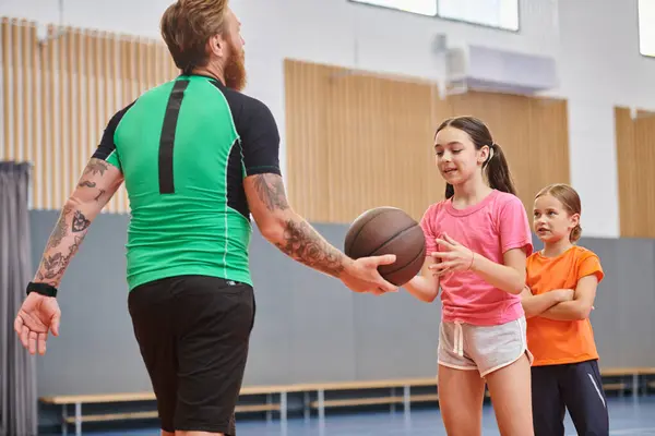 stock image A male teacher stands holding a basketball in front of a diverse group of children in a bright, lively classroom setting.