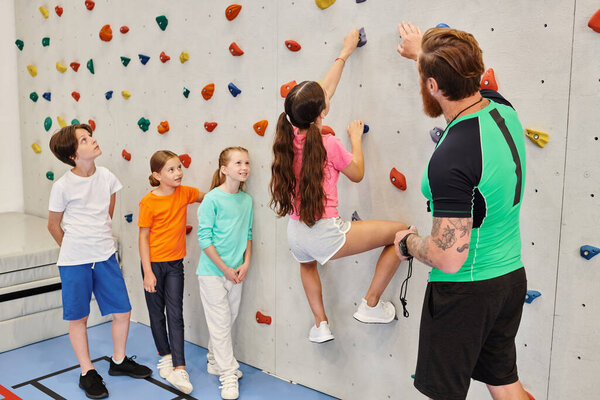 A diverse group of young children stand side by side on a colorful climbing wall, while their teacher guides them through the challenge.
