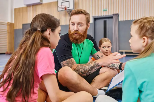stock image A bearded man sits on a basketball court surrounded by diverse children, engaging them in an inspiring lesson about the game.