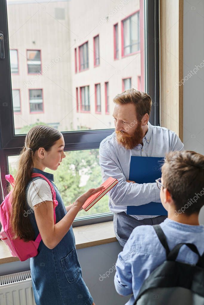 A group of kids eagerly listen as their male teacher instructs them in a bright, lively classroom setting by a large window.