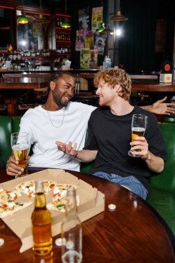 Afro-Amerikan arkadaşıyla barda bira ve pizza içerken el kol hareketi yapan mutlu kızıl adam.
