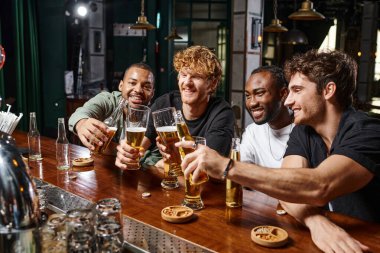 Çok ırklı dört mutlu erkek arkadaş bar tezgahında bir bardak birayla kadeh kaldırıyor.