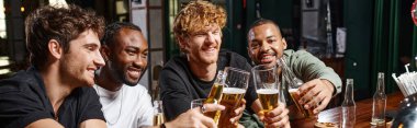 Dört mutlu çok ırklı erkek arkadaş bar tezgahında bir bardak birayla kadeh kaldırıyor.