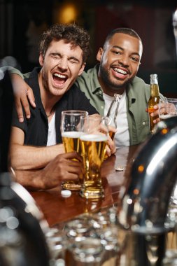 happy african american man hugging excited friend and holding glass of beer in bar, male friendship clipart