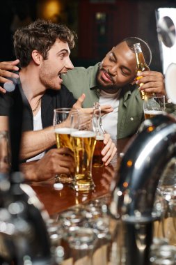 african american man hugging excited friend chatting and holding beer in bar, male friendship clipart