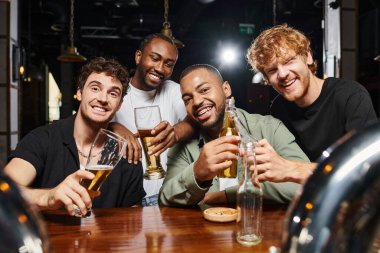 Barda bira bardaklarıyla kameraya bakan dört mutlu çok kültürlü adam, erkek arkadaşlar.