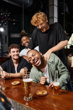 redhead man tickling happy african american friend during bachelor party in bar, male friendship clipart
