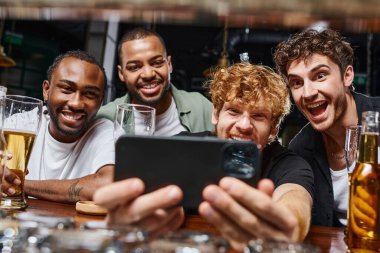 Akıllı telefondan selfie çeken ve bekarlığa veda partisinde bira içen neşeli ırklar arası erkekler.