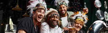 banner, multiethnic male friends in headwear with feathers toasting beer and spending time in bar clipart