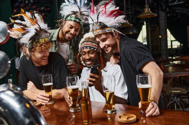 happy multicultural men in headwear with feathers looking at smartphone during bachelor party in bar clipart