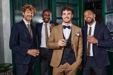 bachelor party, cheerful interracial best men and groom in suits standing with glasses of whiskey clipart