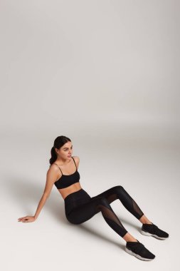 Siyah etekli yorgun sporcu kadın bileğinde spor takip cihazıyla gri arka planda oturuyor.