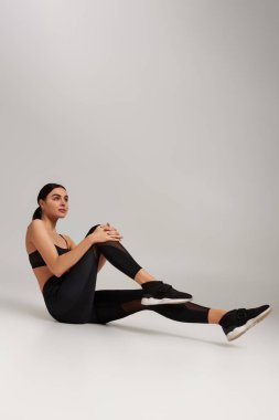 Sporcu kadın aktif giyiniyor. Bileğinde spor takip cihazı var. Gri arka plan üzerinde egzersiz yapıyor.