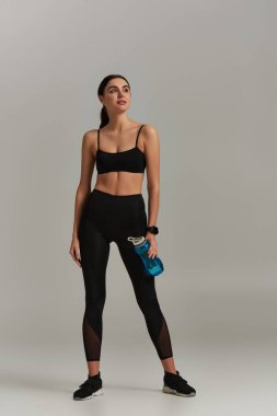 Siyah spor kıyafetli atletik bir kadın gri arka planda dikilirken elinde suyla şişe tutuyor.