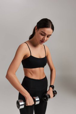 Siyah etekli motive ve güçlü sporcu kadın gri arka planda dambıllarla egzersiz yapıyor.