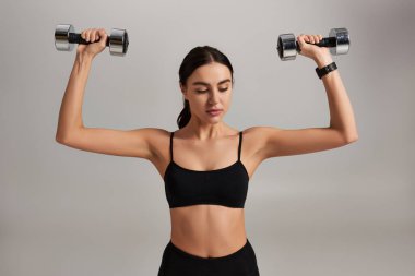 Siyah giyimli genç ve sportif kadın gri arka planda dambıllarla spor yapıyor.