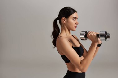 Siyah etekli spor giyen genç bayanın yan görünüşü gri arka planda dambılları kaldırıyor