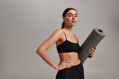 Sporcu kadın spor kıyafetlerini tutuyor ve elleri kalçasında gri arka planda duruyor.