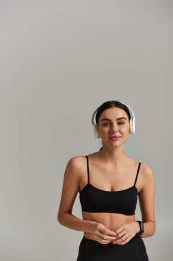 Gri arka planda kablosuz kulaklıklarla müzik dinleyen ekinlerin içinde çekici bir sporcu kadın.