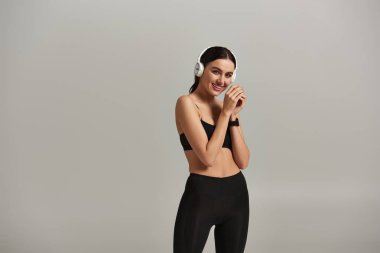 Aktif çalışan memnun sporcu kadın gri arka planda kablosuz kulaklıkla müzik dinliyor.