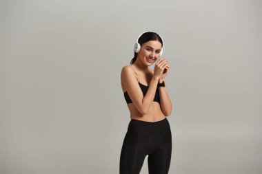 pleased sportswoman in active wear listening music in wireless headphones on grey background clipart