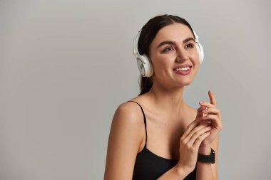 Aktif çalışan mutlu genç kadın gri arka planda kablosuz kulaklıkla müzik dinliyor.