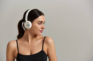 Aktif çalışan güzel genç bir kadın gri arka planda kablosuz kulaklıkla müzik dinliyor.
