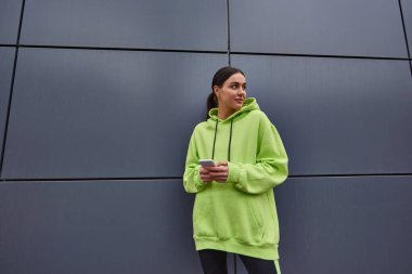 Limon rengi kapüşonlu neşeli kadın elinde akıllı bir telefonla gri duvarın yanında dikilirken yüzünü çeviriyor.