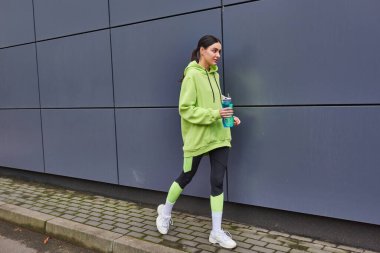 Kapüşonlu ve taytlı çekici bir sporcu. Gri duvarın yanında bir şişe suyla yürüyor.