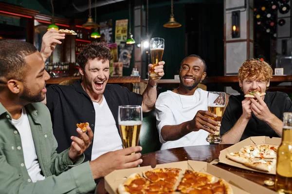 excited man holding pizza and beer while screaming near interracial friends in bar, male friendship