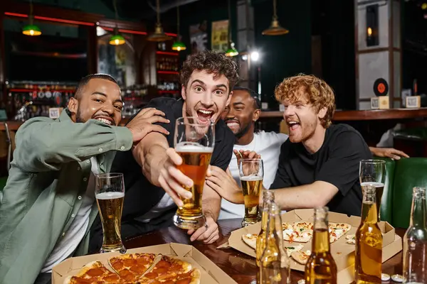 group of excited multicultural friends toasting with glasses of beer in bar, men on bachelor party