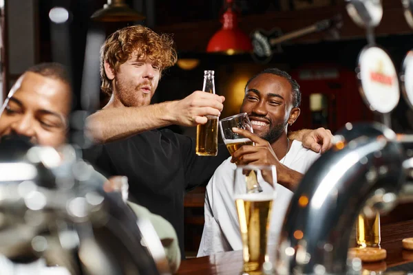 cheerful multiethnic friends hugging and toasting glasses of beer while spending time in bar