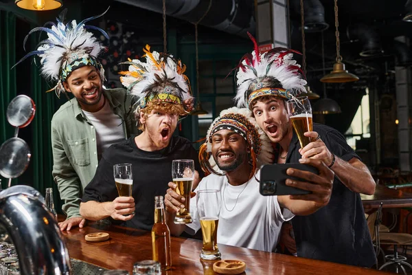 stock image happy multicultural men in headwear with feathers taking selfie on smartphone during bachelor party