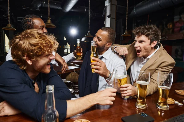 emotional and interracial colleagues in formal wear drinking beer in bar, having fun after work