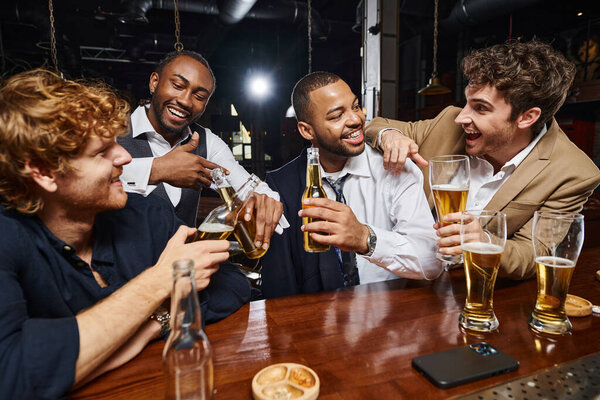 cheerful interracial colleagues in formal wear toasting beer in bar, men having fun after work