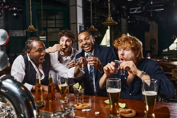 group of four happy and drunk interracial men in formal wear drinking tequila in bar after work