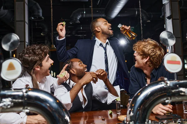 happy men looking at african american friend drinking beer from two bottles after work in bar
