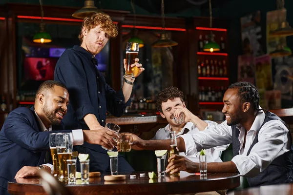 cheerful and drunk multiethnic colleagues toasting with glasses of whiskey after work in bar