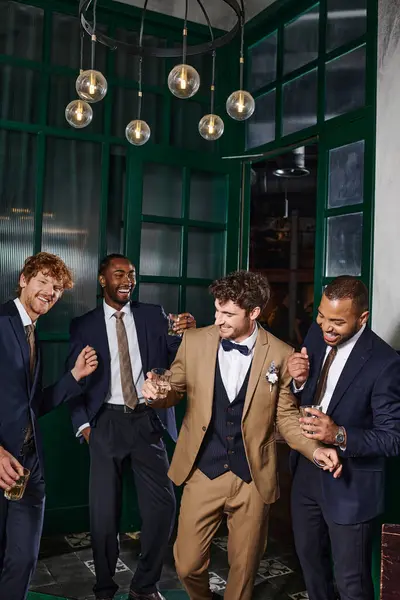 bachelor party, interracial best men and groom laughing while standing with glasses of whiskey