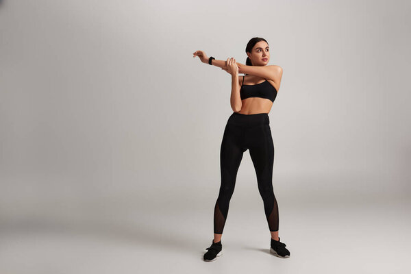 young sporty woman in black sportswear with fitness tracker on wrist stretching on grey backdrop