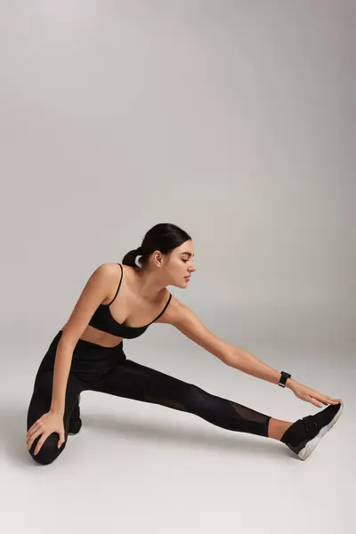 motivated woman in black active wear with fitness tracker on wrist stretching leg on grey backdrop