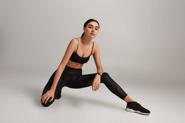 beautiful woman in black active wear with fitness tracker on wrist stretching leg on grey backdrop
