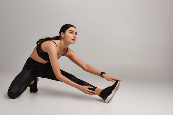 young sportswoman in black active wear with fitness tracker on wrist stretching leg on grey backdrop