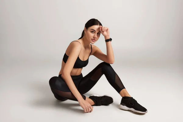 brunette sportswoman in black active wear with fitness tracker on wrist sitting on grey backdrop