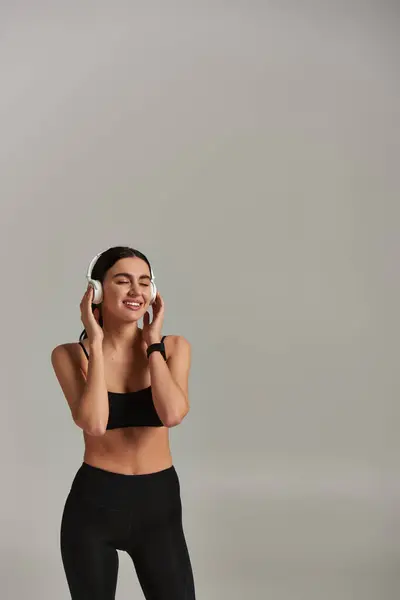 cheerful sportswoman in active wear listening music in wireless headphones on grey background