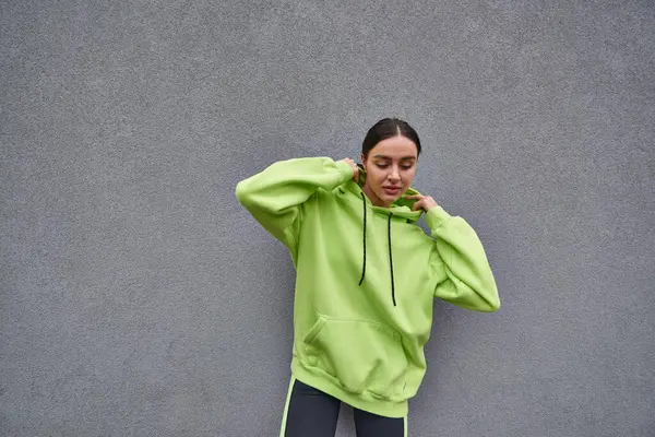 brunette woman in trendy lime color hoodie adjusting hood and standing near grey concrete wall