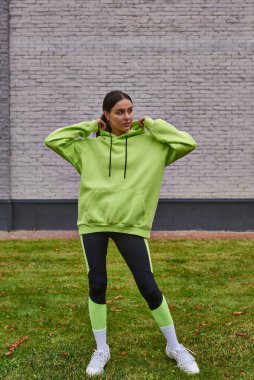 stylish sportswoman in lime color hoodie and leggings standing on green grass, healthy lifestyle clipart