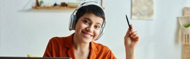 jolly young student with headphones and piercing smiling while studying at home, education, banner clipart