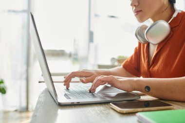 Kırpılmış turuncu gömlekli, kulaklıklı, dövmeli, laptopta eğitim gören genç kadın görüntüsü.