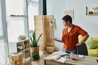 Çok çalışan kısa saçlı öğrenci masasının yanındaki odada poz veriyor ve elinde dizüstü bilgisayar ve eğitim var.